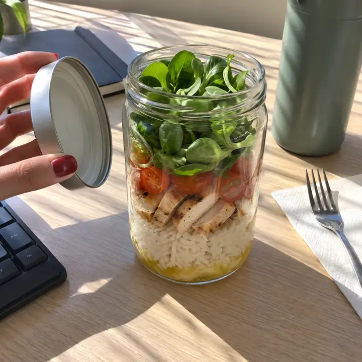 Tarro de arroz con pollo, tomate cherry y canónigos arriba, recién abierto sobre una mesa de oficina