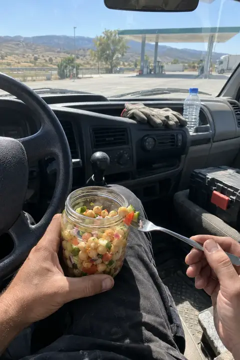 Tarro de ensalada de legumbres apoyado en la rodilla, dentro de una furgoneta de trabajo en un área de servicio